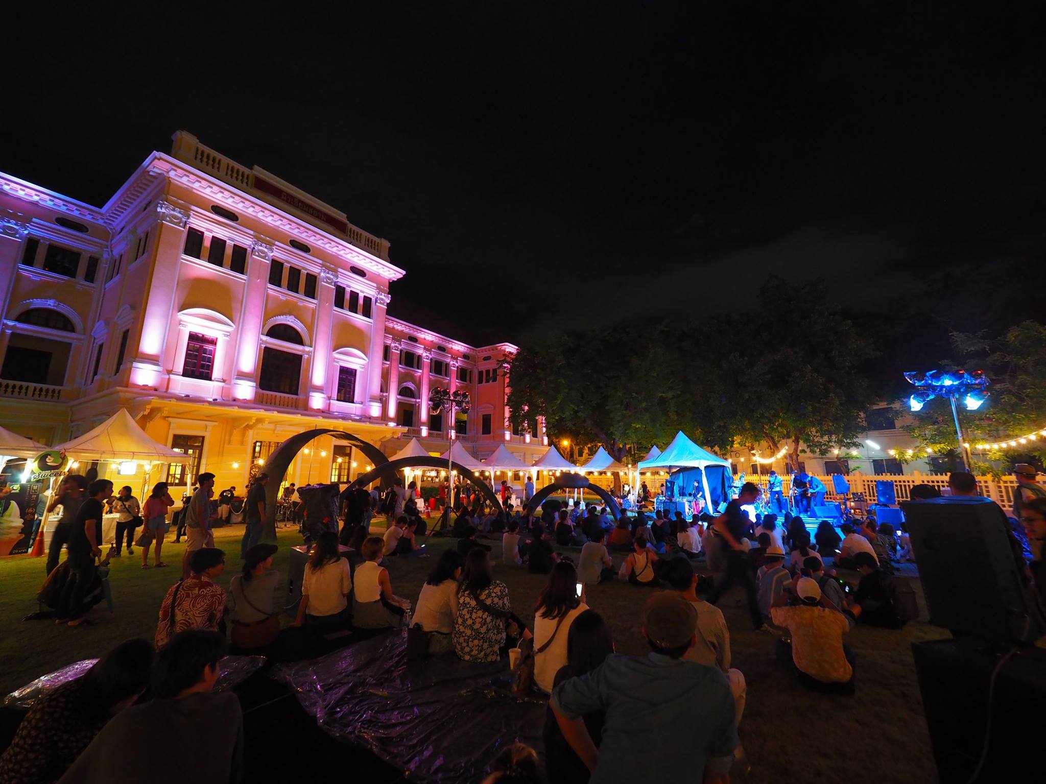 The best museums and galleries in Bangkok | a view of a sparse crowd sat on the floor, cross legged, outside the Museum Siam at night. The museum is illuminated by pink lighting, and a band perform on a stage in the distance.