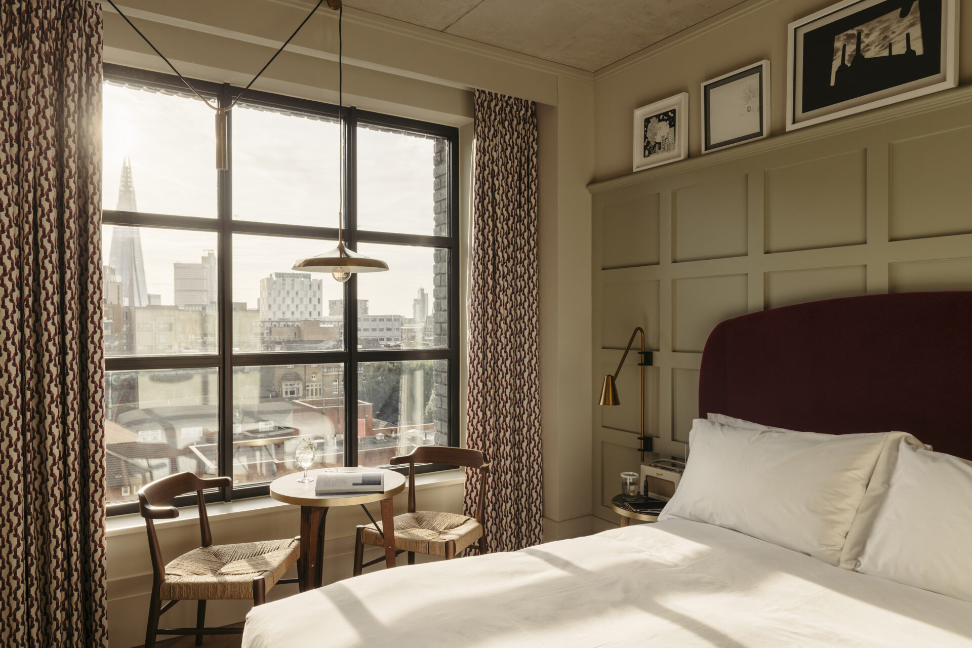 Best hotels in Shoreditch, Dalston and Hackney | A bedroom view inside The Hoxton Hotel in Shoreditch. A large window shines light over a table and chairs and a bed, and painted wood panelling decorates the wall behind the bed. Through the window, The Shard can be seen in the distance.