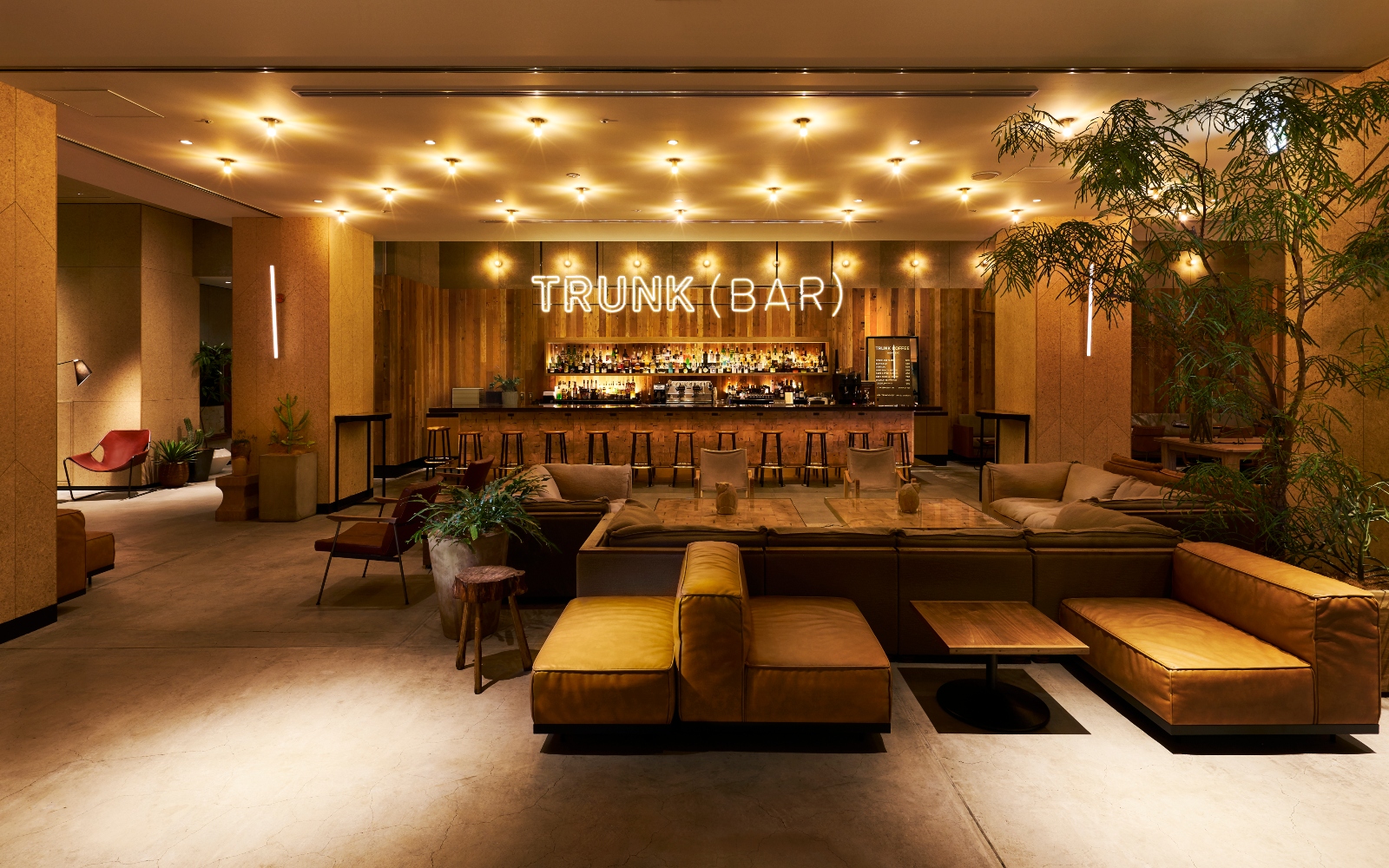 The communal bar area at Shibuya Trunk hotel Tokyo, with low wooden furniture, mustard-yellow leather sofas and oriental-inspired greenery, in front of the long bar