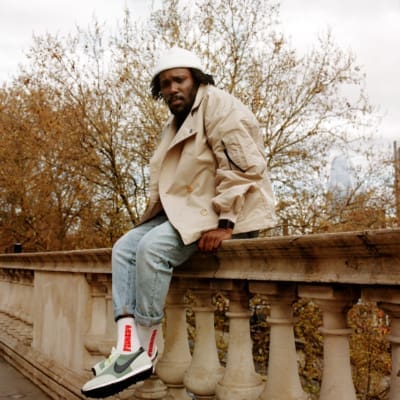 Akwasi Brenya-Mensa | Akwasi wearing jeans, white trainers, a white beanie and a trench coat sat on a rail at Somerset House