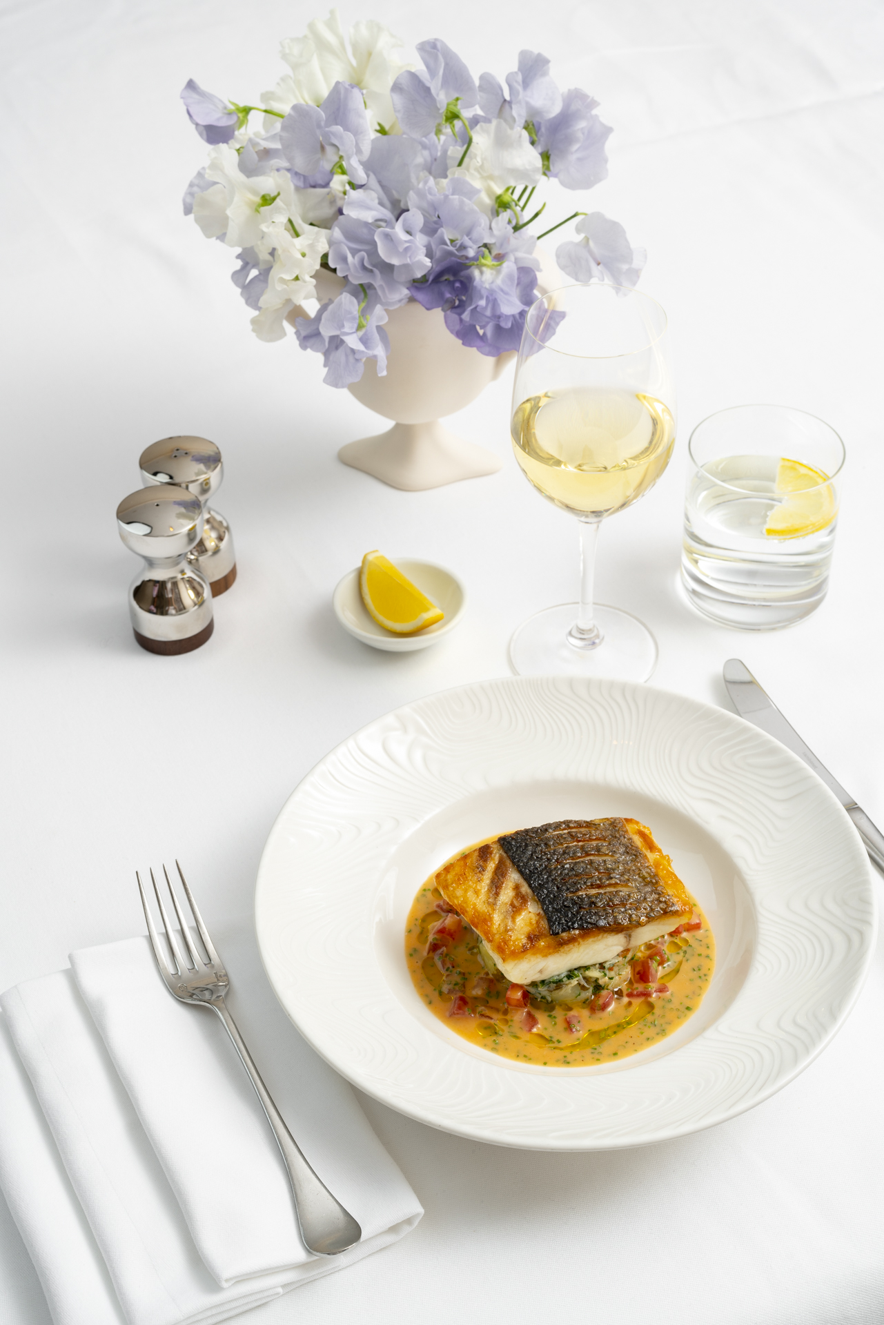 Things to do in London in June | a tranche of trout on a bed of vegetables in a white bowl on a white tablecloth with silverware, silver salt and pepper shakers and a bouquet of purple flowers at Sotheby's
