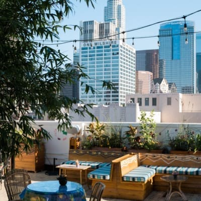 Best rooftop bars in LA | a tree, some wooden bench seating with blue striped cushions and tables at rooftop bar the Broken Shaker at the Freehand Hotel, with a backdrop of blue sky and skyscrapers