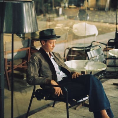 An interview with Thai pop star Hugo Chakrabongse | Hugo wearing blue bell-bottomed jeans, a white shirt, grey jacket and wide-brimmed hat on a metal-framed chair in front of floor-to-ceiling windows