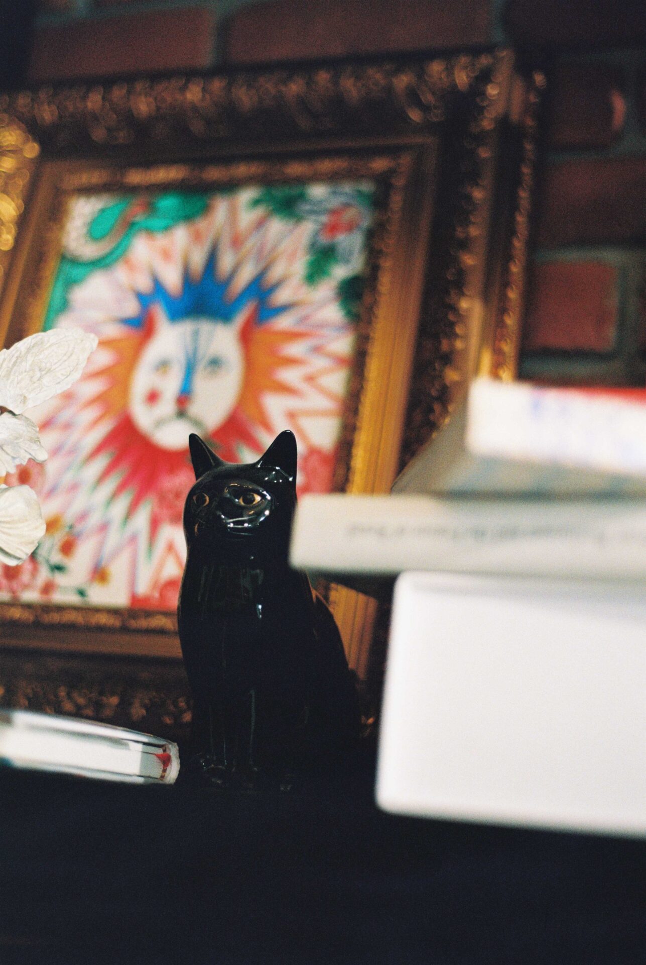 Phannapast | A small statue of a black cat sits on a shelf in front of a colourful line drawing of a lion inside a gold frame