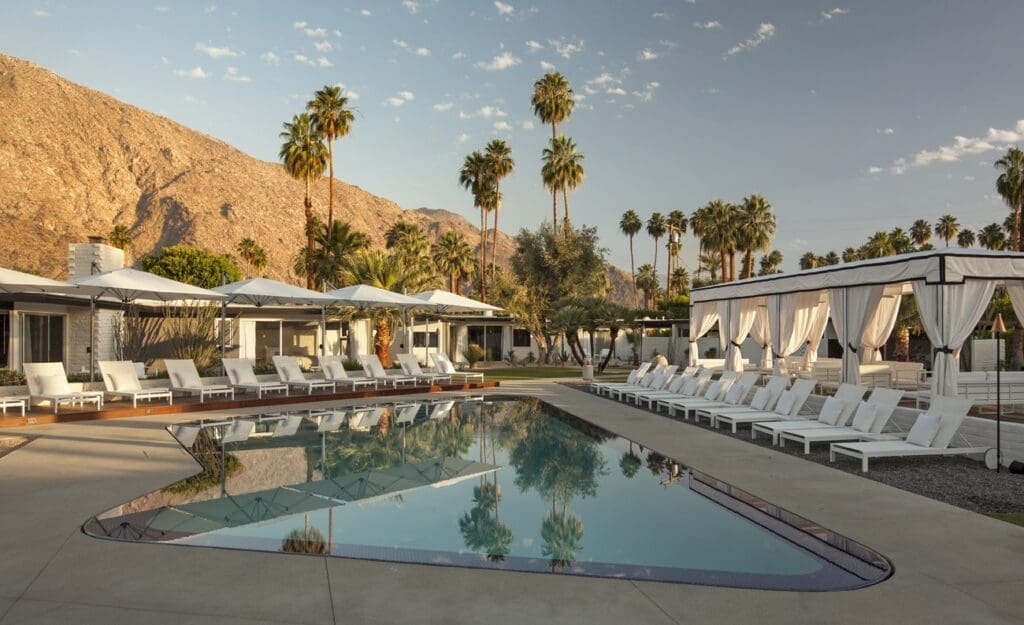 What to do in Palm Springs, California | A pool surrounded by white sun beds and cabanas in front of a desert mountain backdrop with palm trees