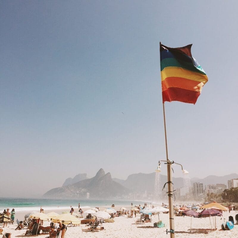 A pride flag sits in the sand on a busy beach