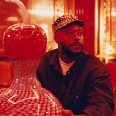 A portrait of Yinka Ilori sat with his original Murano glass sculpture in the red-lit interiors of Mayfair private member's club George