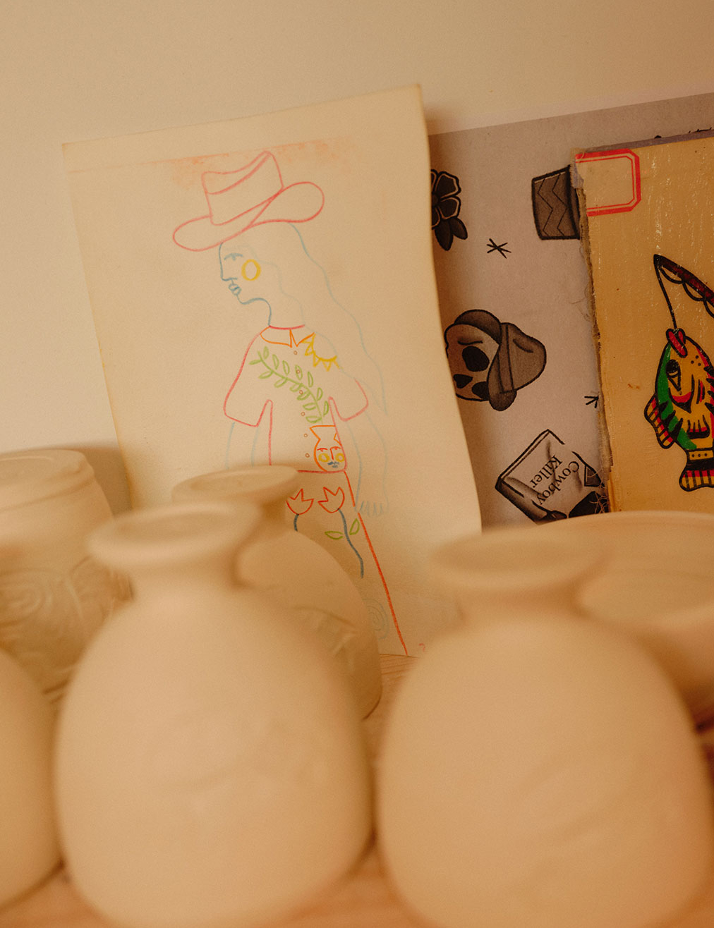 Unpainted vases resting in the studio with illustration ideas sketched on paper behind.