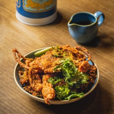 Soft shell crab served in a bowl on a wooden tabletop next to a small blue jug, at Kotuwa restaurant in Singapore.