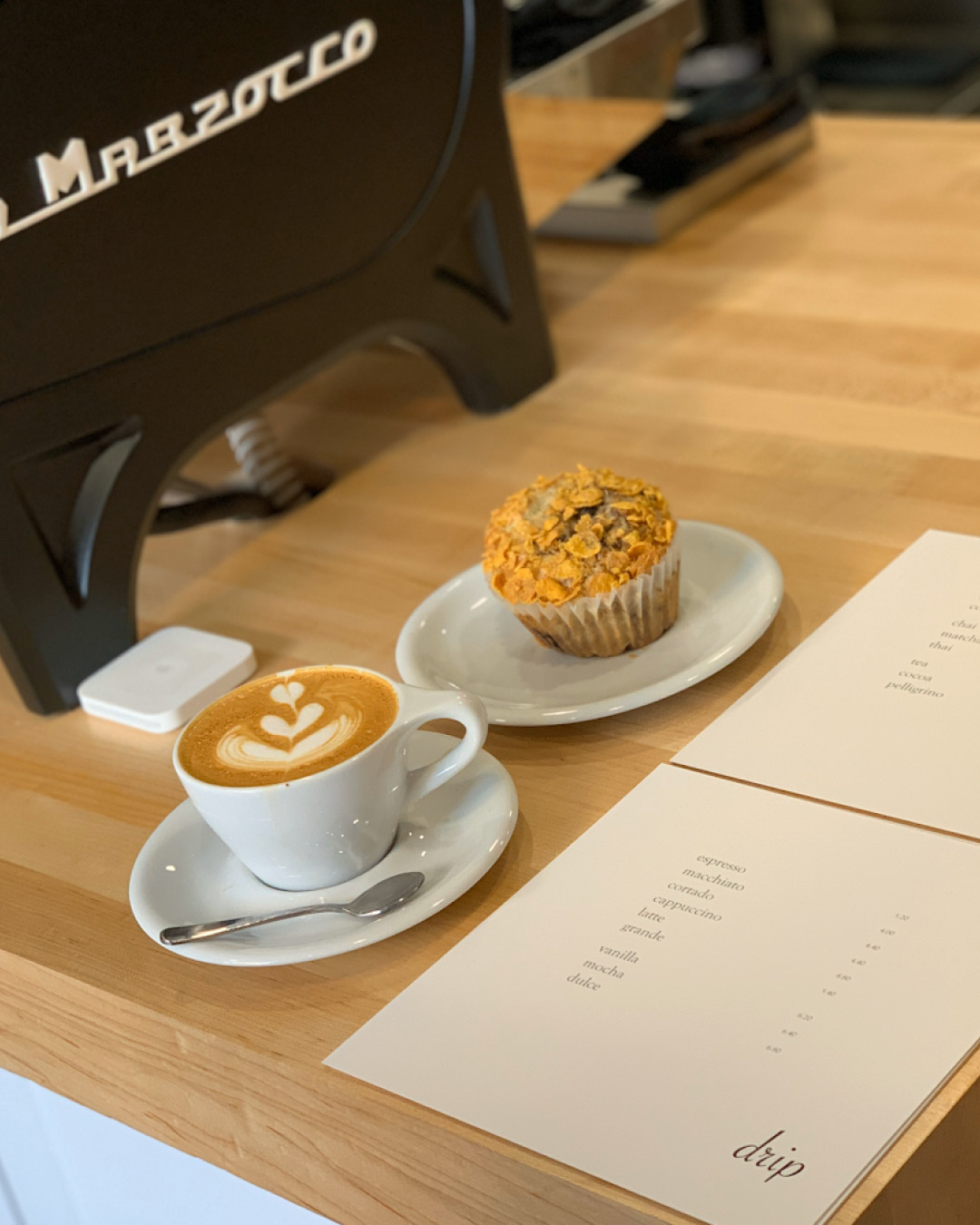 A latte and cornflake muffin served at Drip Coffee Makers on a wooden countertop next to printed menus and a black coffee machine.