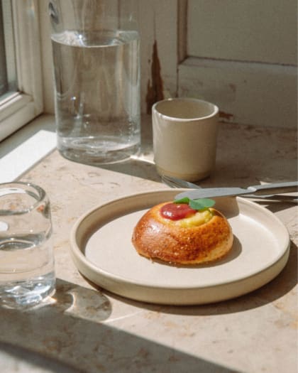 A sourdough bun on a beige plate, with a glass of water, a mug, and a jug on a marble windowsill.