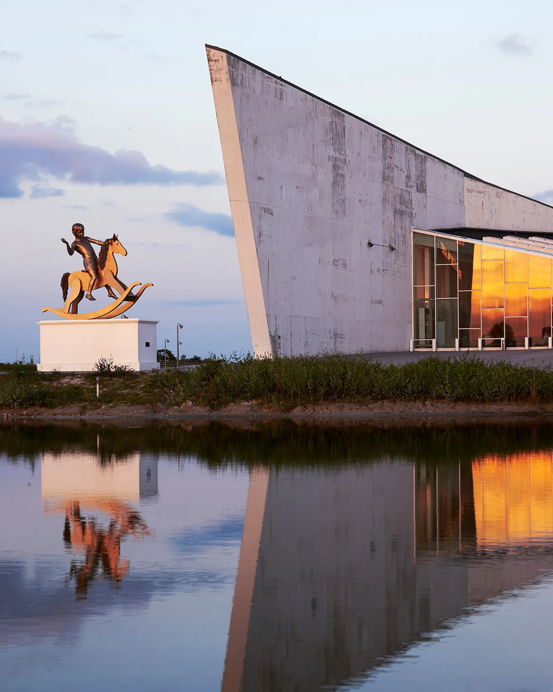 The angular facade of ARKEN Museum of Contemporary Art, reflected in water, with a sculpture outside.