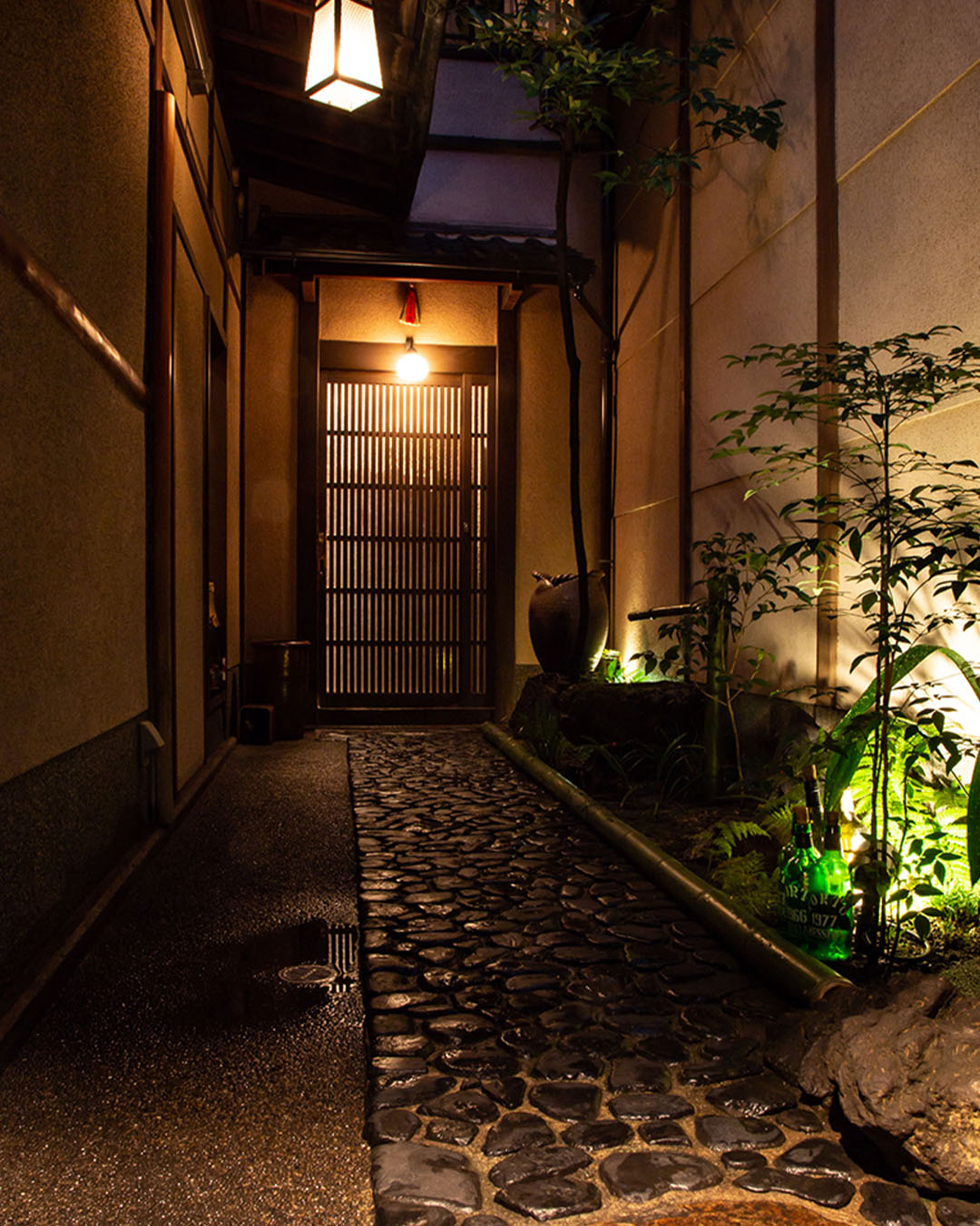 An alleyway entrance to Rocking Chair bar in Kyoto, where a cobbled walkway leads to a reed door flanked by ceramics and a tall tree.