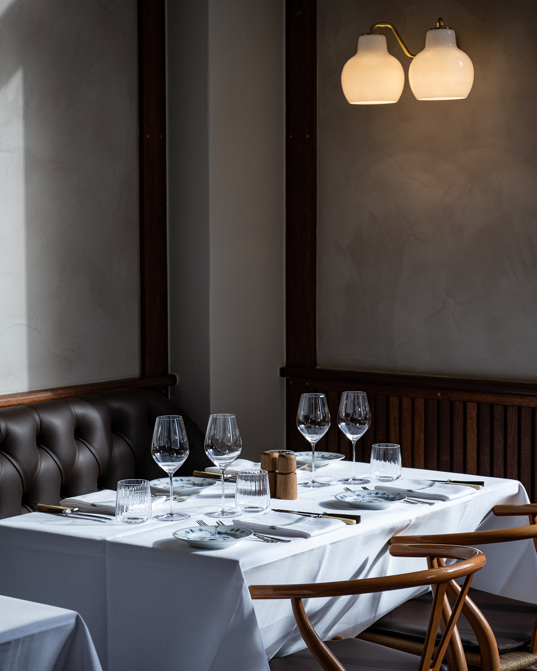 Table setting at Restaurant Møntergade: a white tablecloth drapes over a table, surrounded by a banquet and two wooden chairs, with wine glasses, cutlery and napkins arranged on the table.