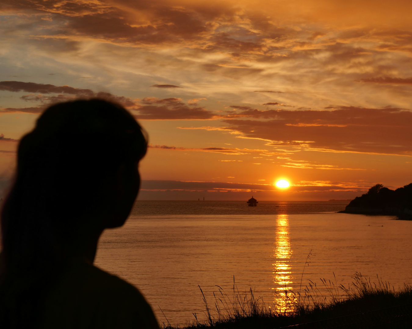 A silhouette of a woman's head looks out to the sea, where an orange sun is setting in a cloudy sky.