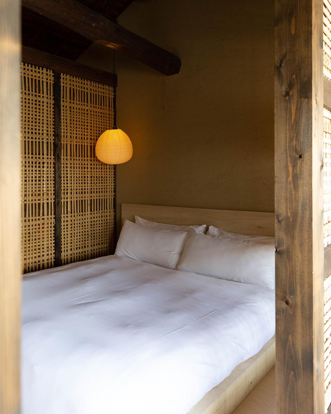 A shoji paper lamp hangs over a double bed made up with white sheets in a small wooden room with a woven screen and wood panelling at Maana Kiyomizu in Kyoto.