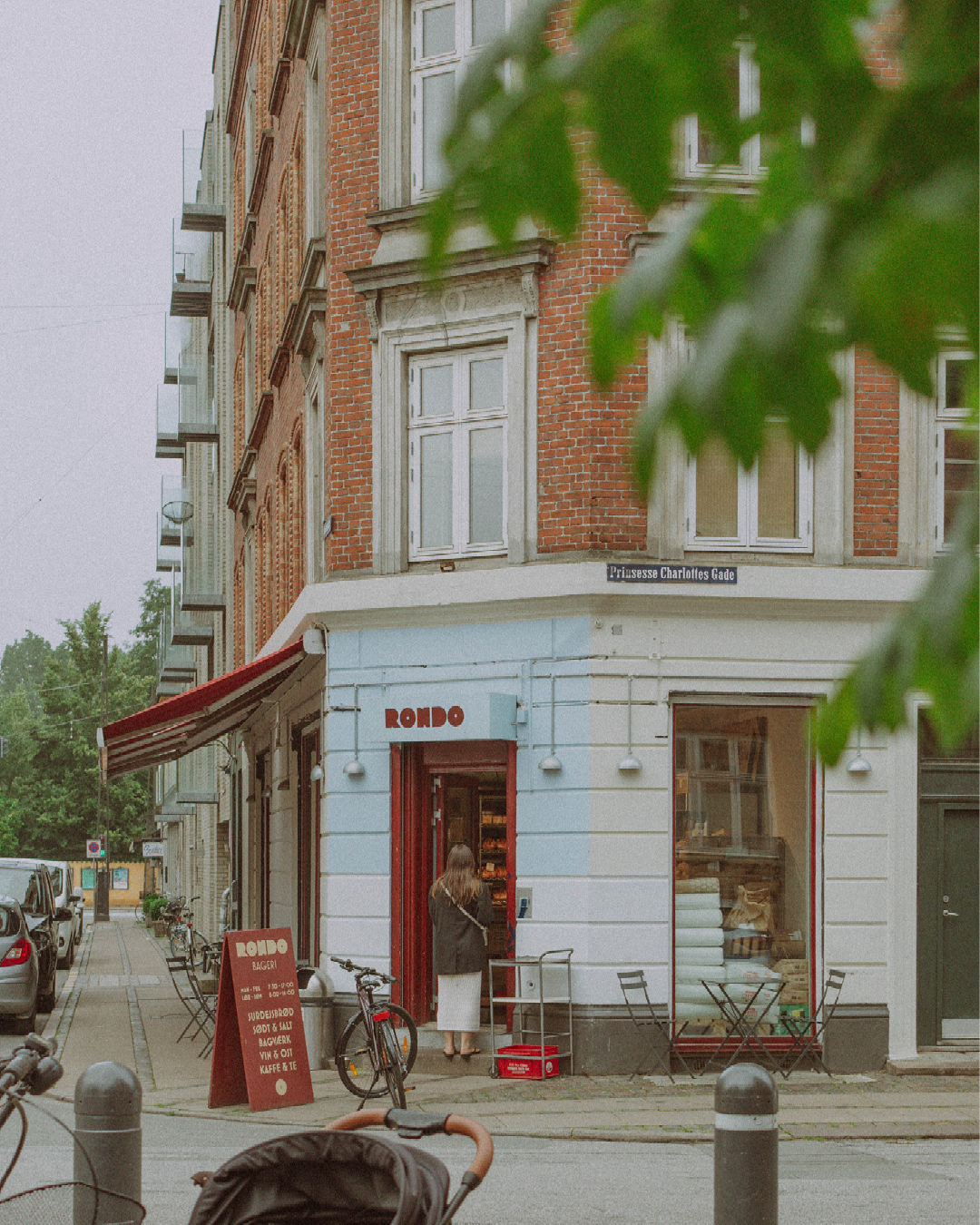 A street view of Rondo Bakery on the corner of two streets. Its exterior walls are painted white, and its doorway and name are painted in red, with a red awning to the left.