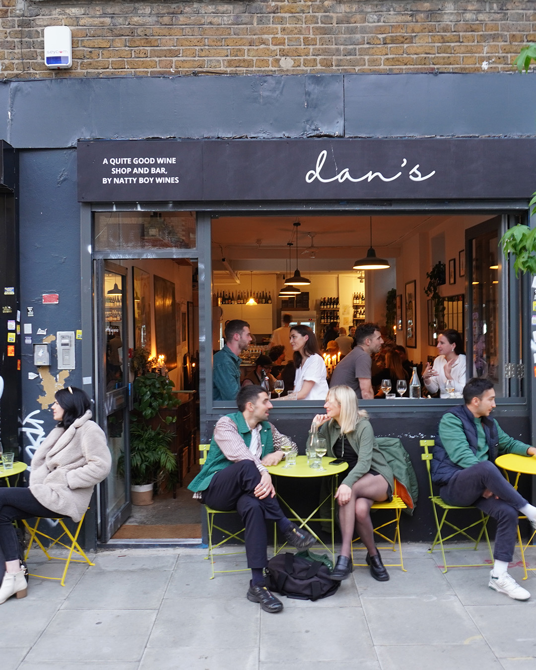 The exterior of Dan's wine bar in Dalston, with customers sitting on yellow chairs